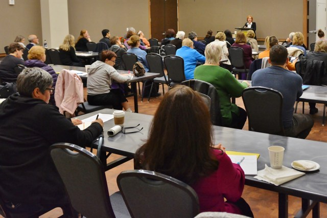 A classroom full of people look at a presenter standing at a podium